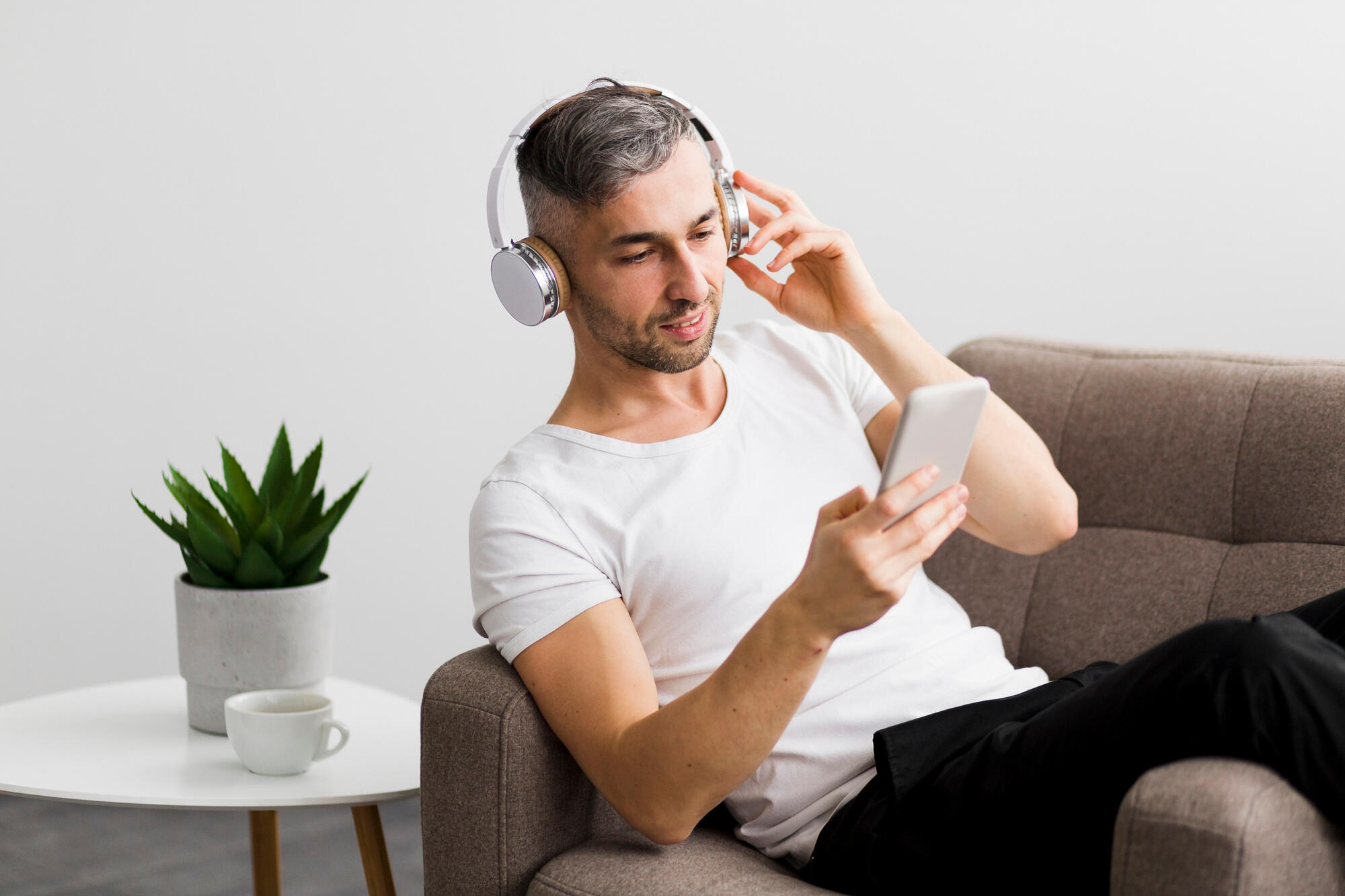 Man wearing headphones, sitting on a couch and holding a smartphone, with a small table holding a plant and a cup nearby.