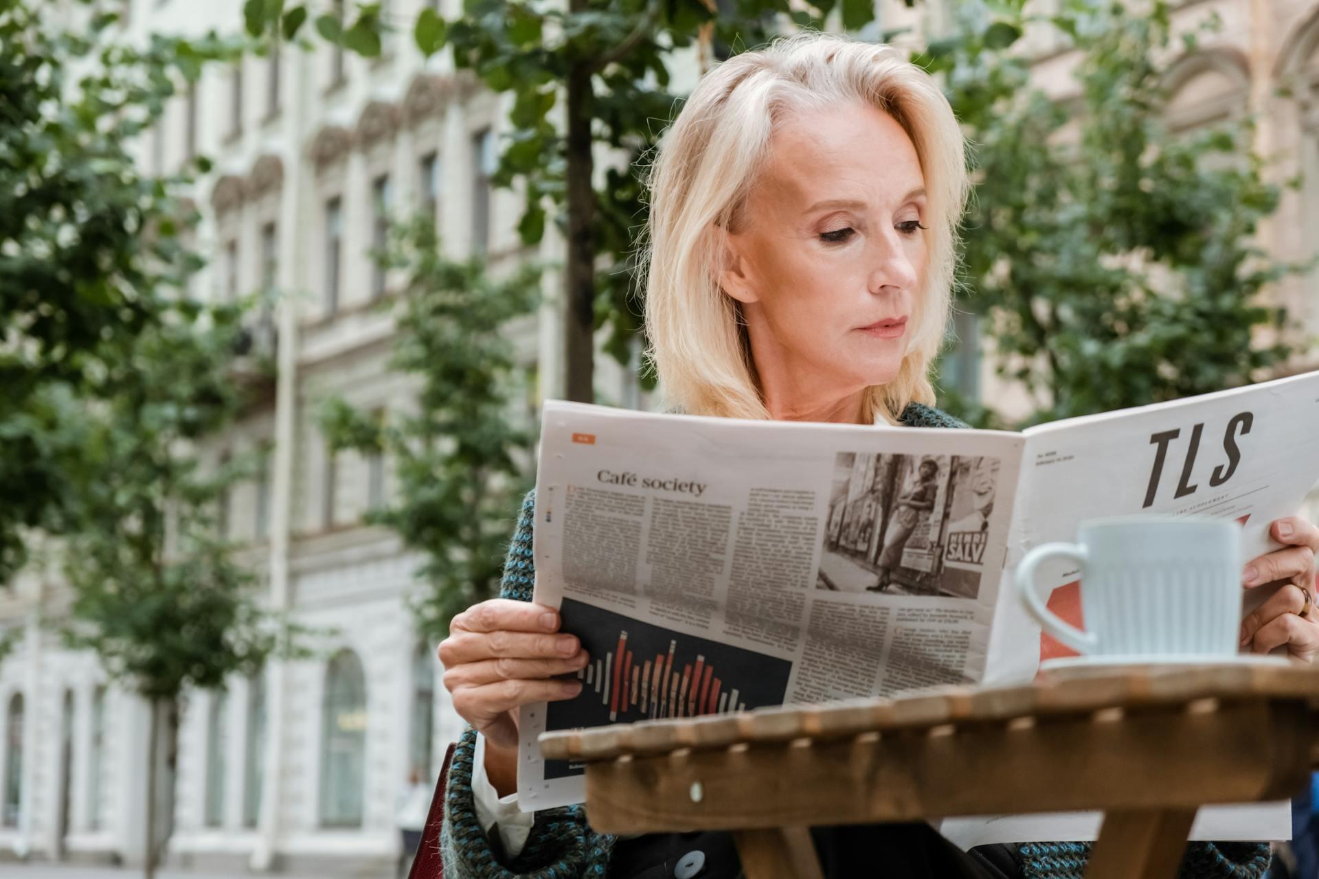 Person sitting outdoors at a wooden table, reading a newspaper with a white cup nearby and trees in the background.