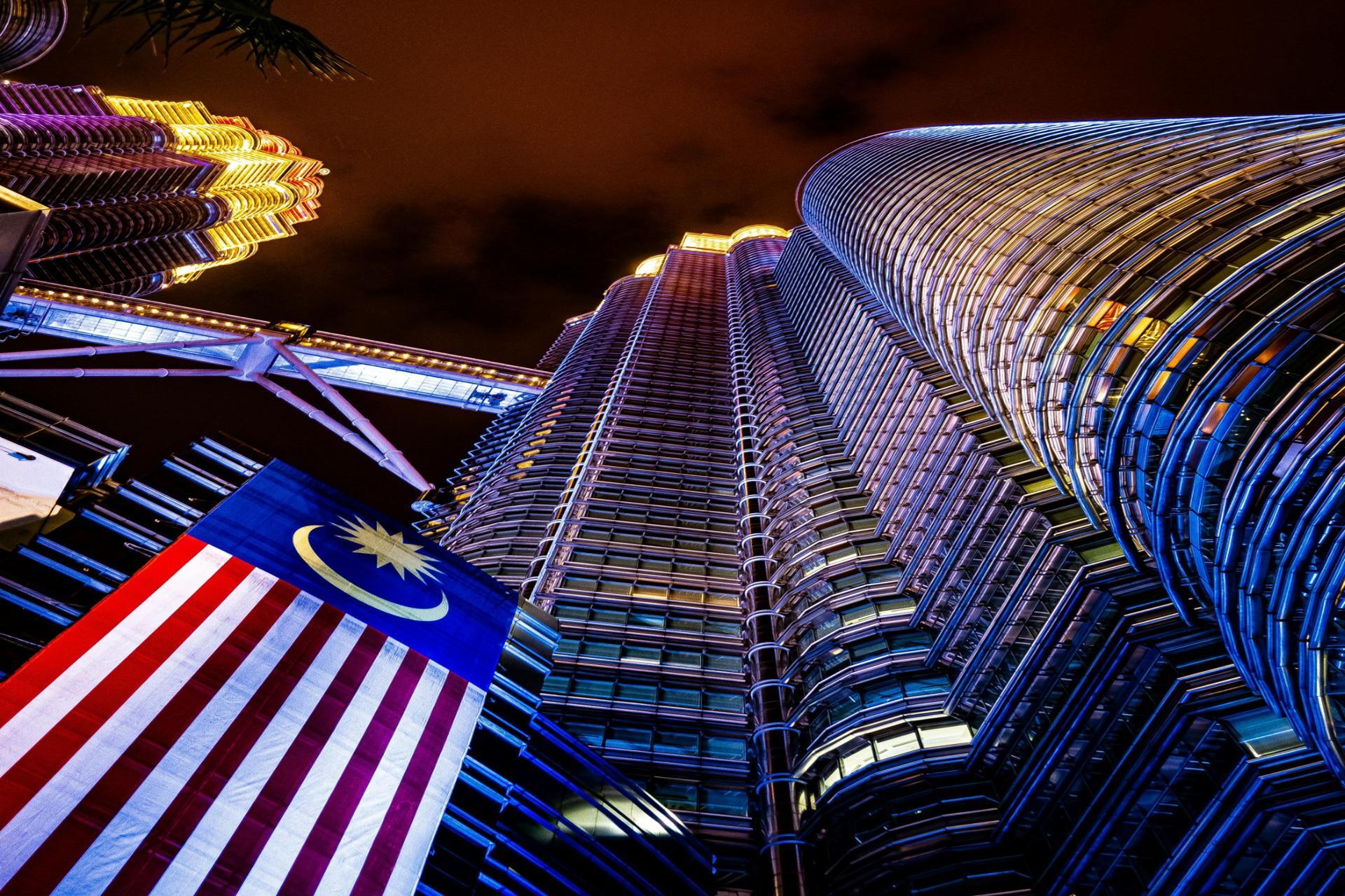Kuala Lumpur city skyline at night featuring the illuminated Petronas Twin Towers with modern buildings and the Malaysian flag in the foreground