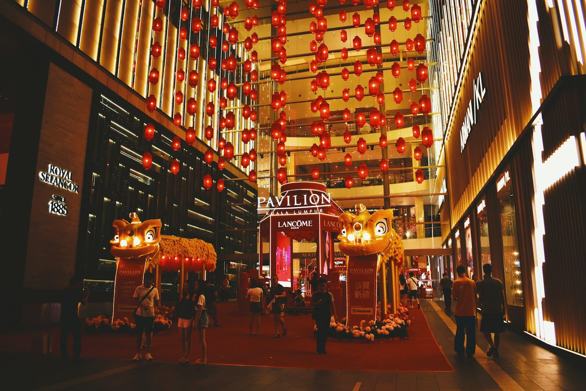 Shopping mall entrance adorned with vibrant red lanterns and two lion statues for Lunar New Year. Warm lighting and festive atmosphere with people walking.