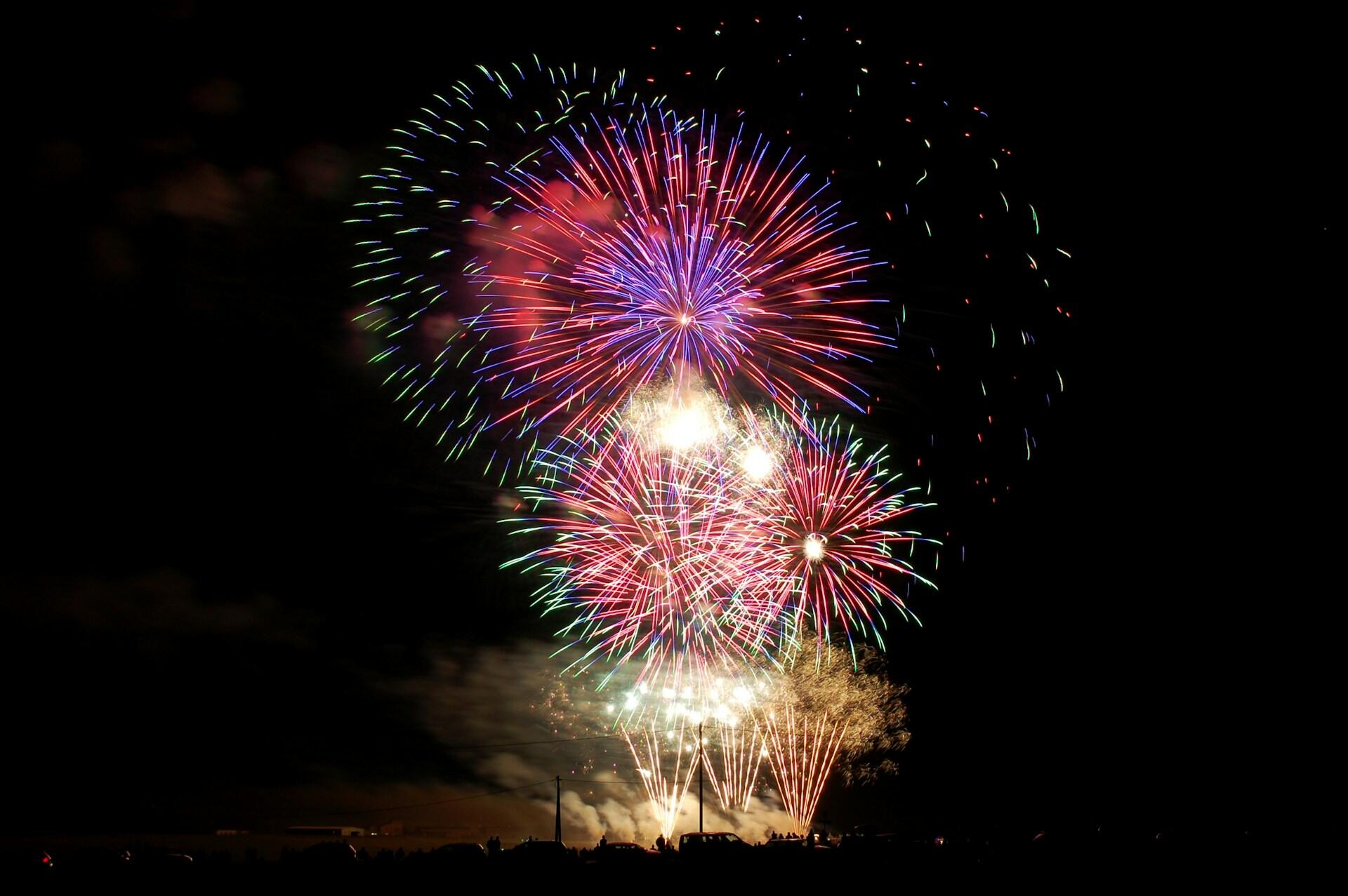 Vibrant fireworks burst in a dark sky, with red, blue, and green hues illuminating the night. A large crowd silhouettes below, creating a festive atmosphere.