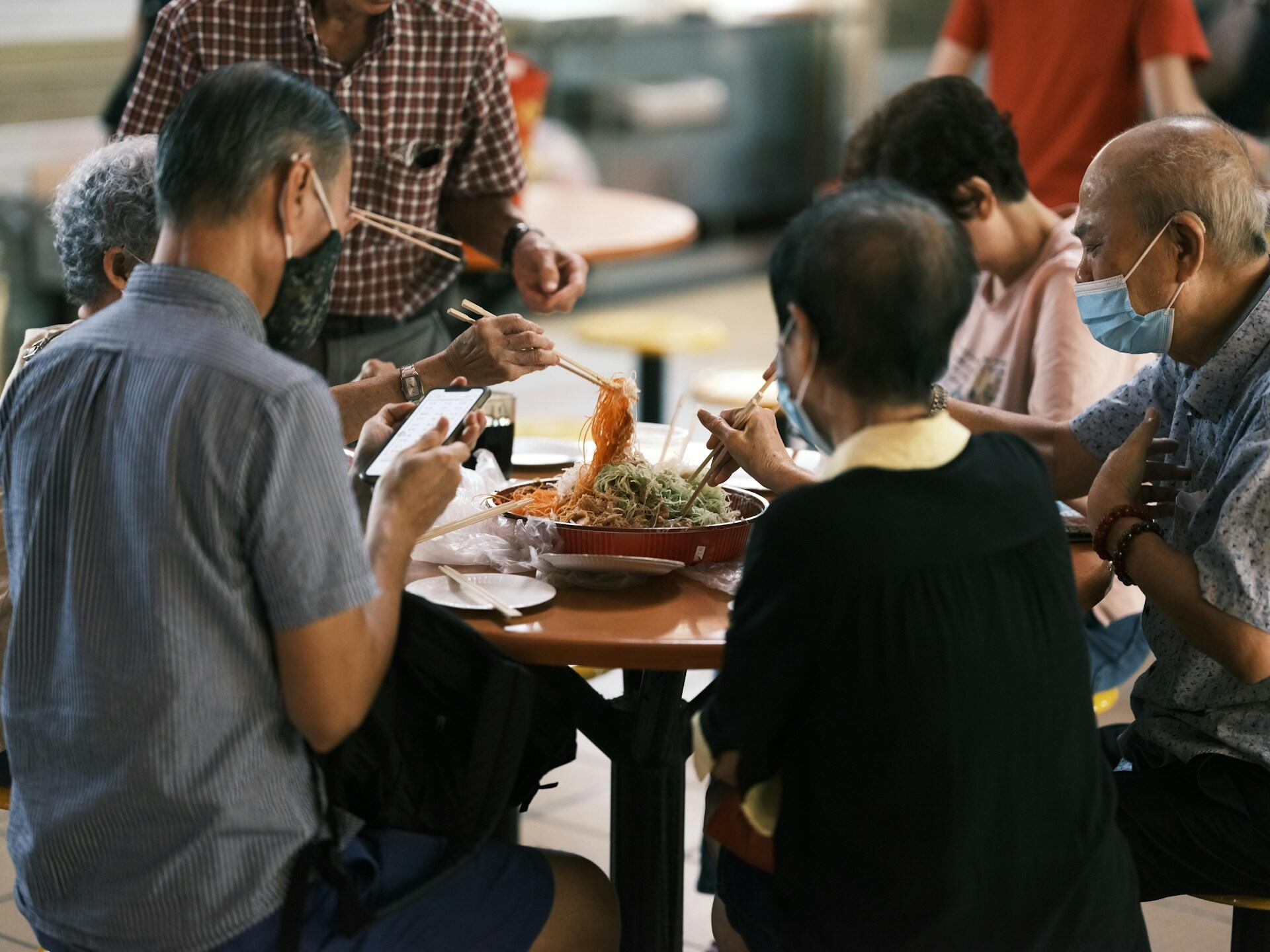 A group of six older adults tossing Yee Sang (prosperity toss) using chopsticks as high as possible. 