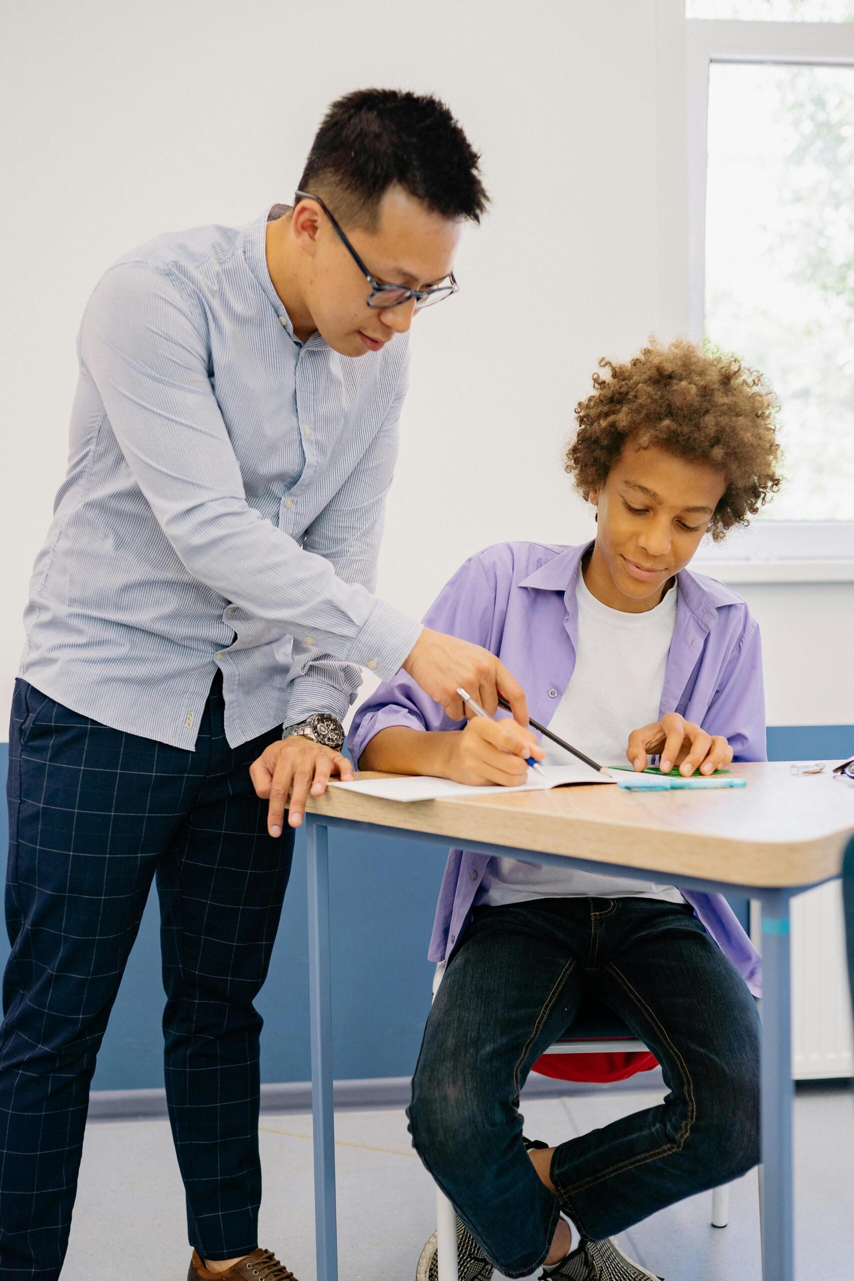 A male teacher guiding a student through homework in a classroom setting.