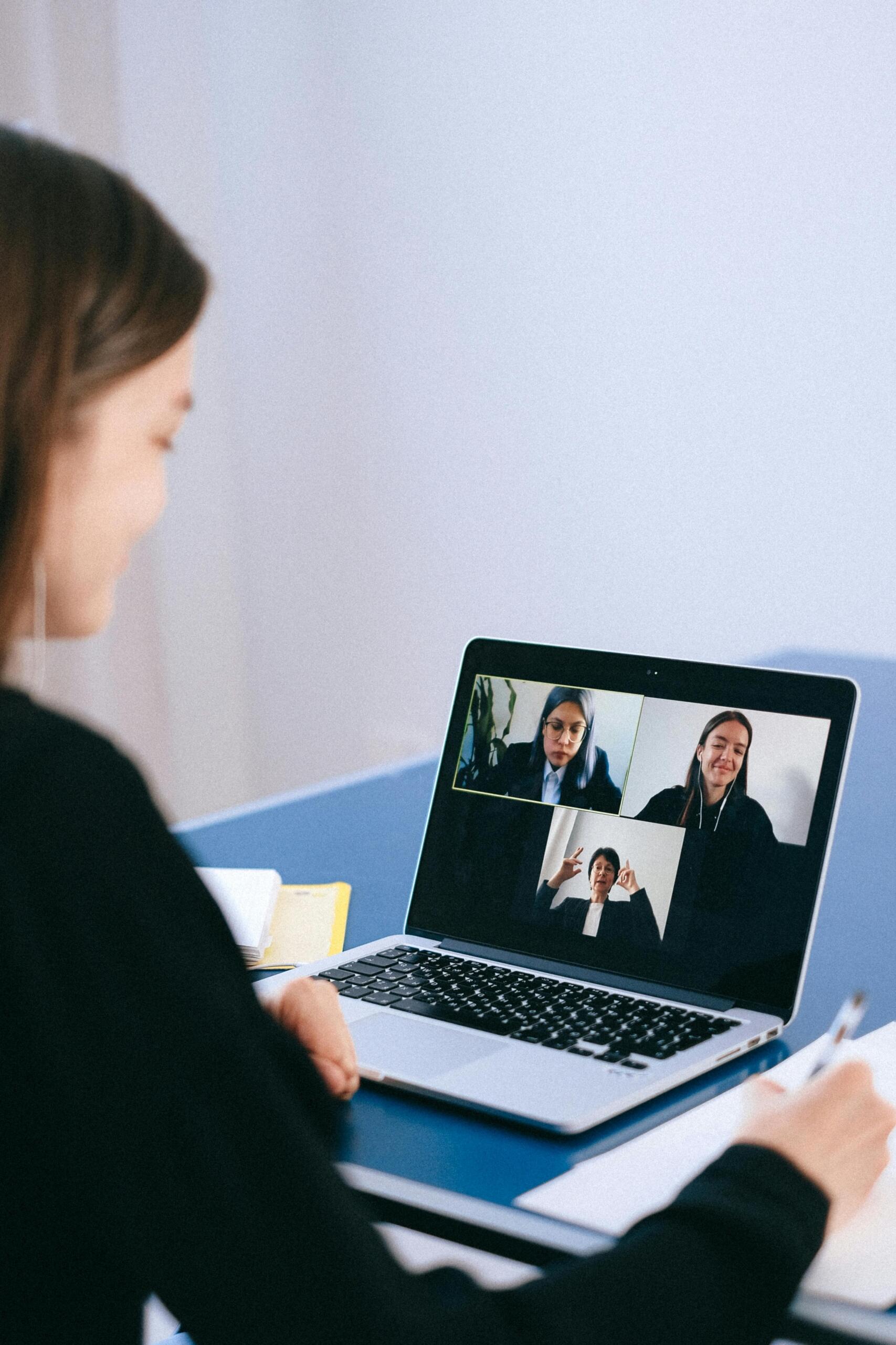 A woman at a desk using a laptop, with two people visible on the screen during a video call.