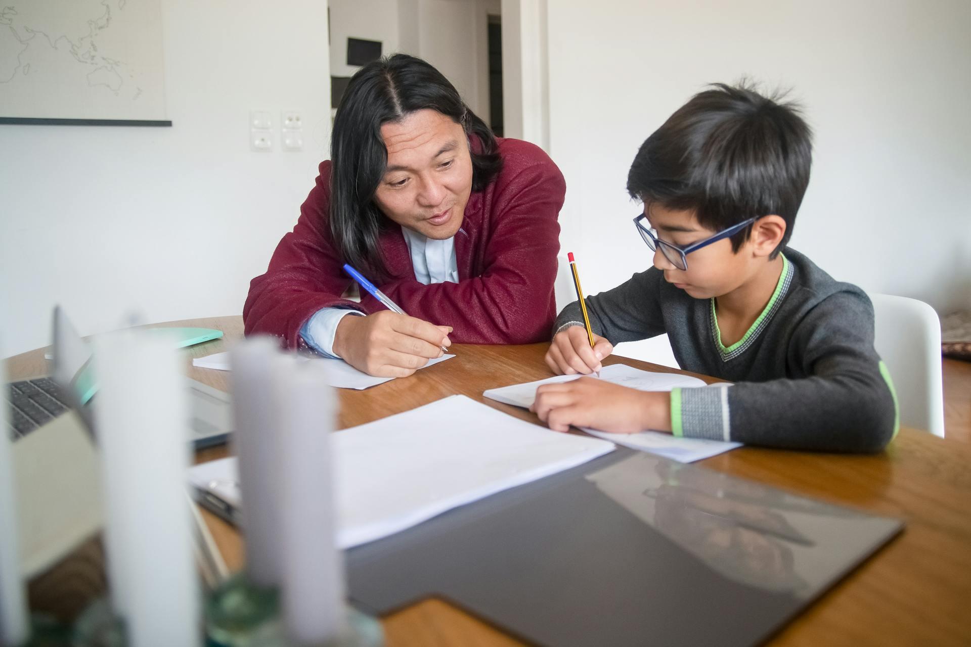 An adult and a child sit at a table working together, both writing on paper with pencils, surrounded by notebooks and a laptop.