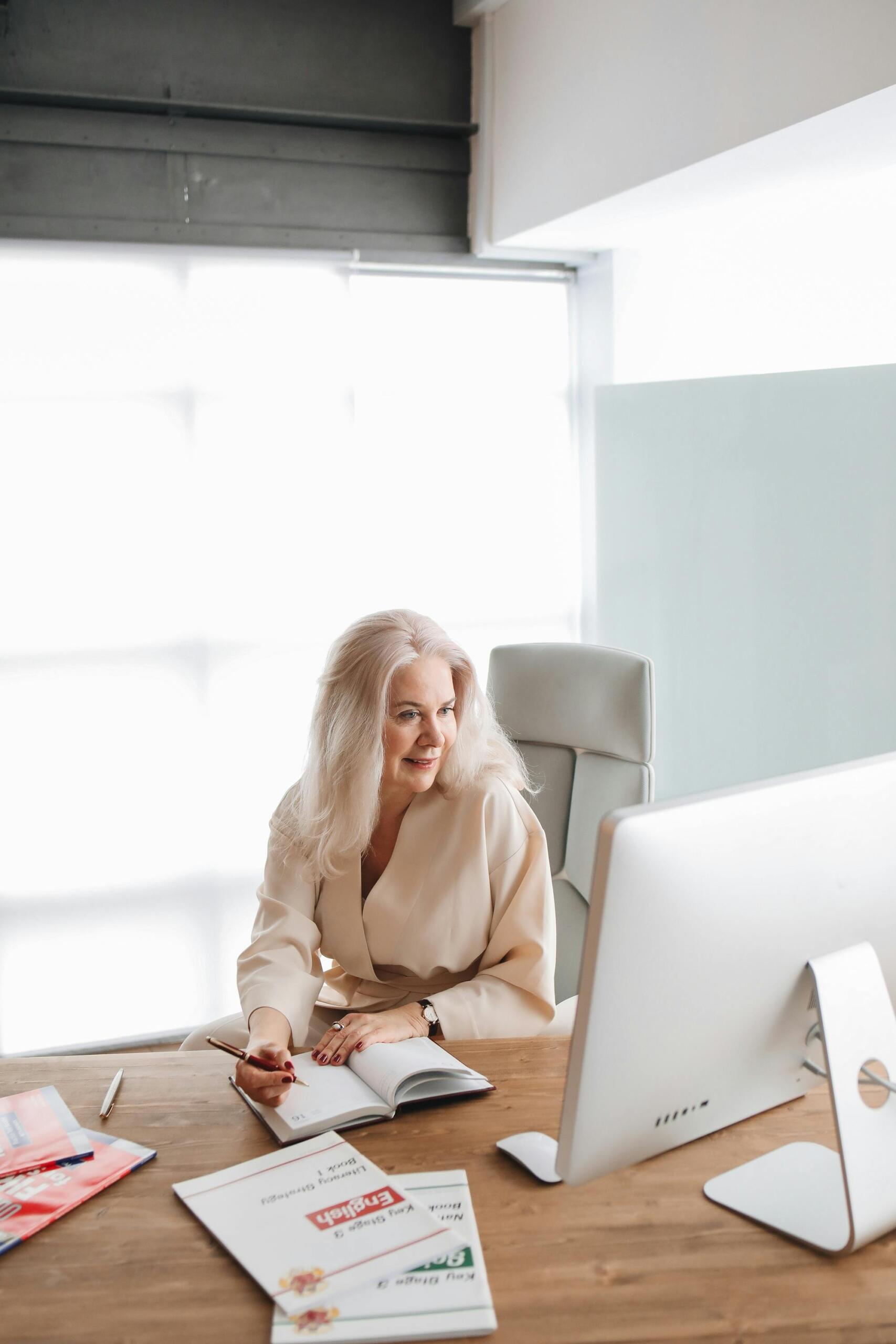A woman at her desk full of office supplies, writing in her notebook in front of her desktop.