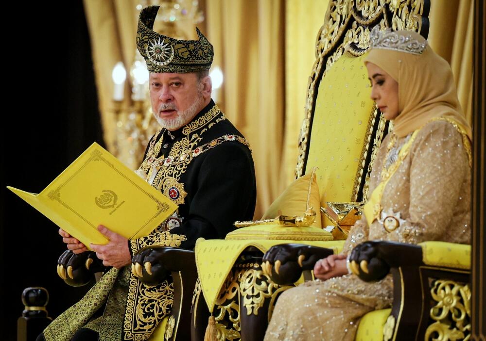 Malaysia's current king, Sultan Ibrahim in ceremonial attire reading a document beside Raja Permaisuri Agong in a gold gown seated on a throne.