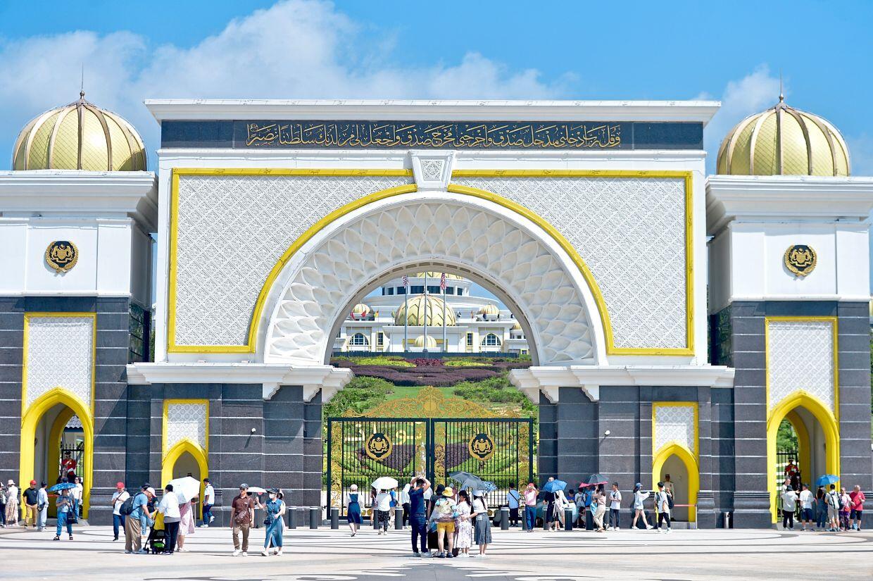 A large crowd gathers outside the ornate main gate of Istana Negara, the Malaysian National Palace, with domes and Islamic script visible under a blue sky.