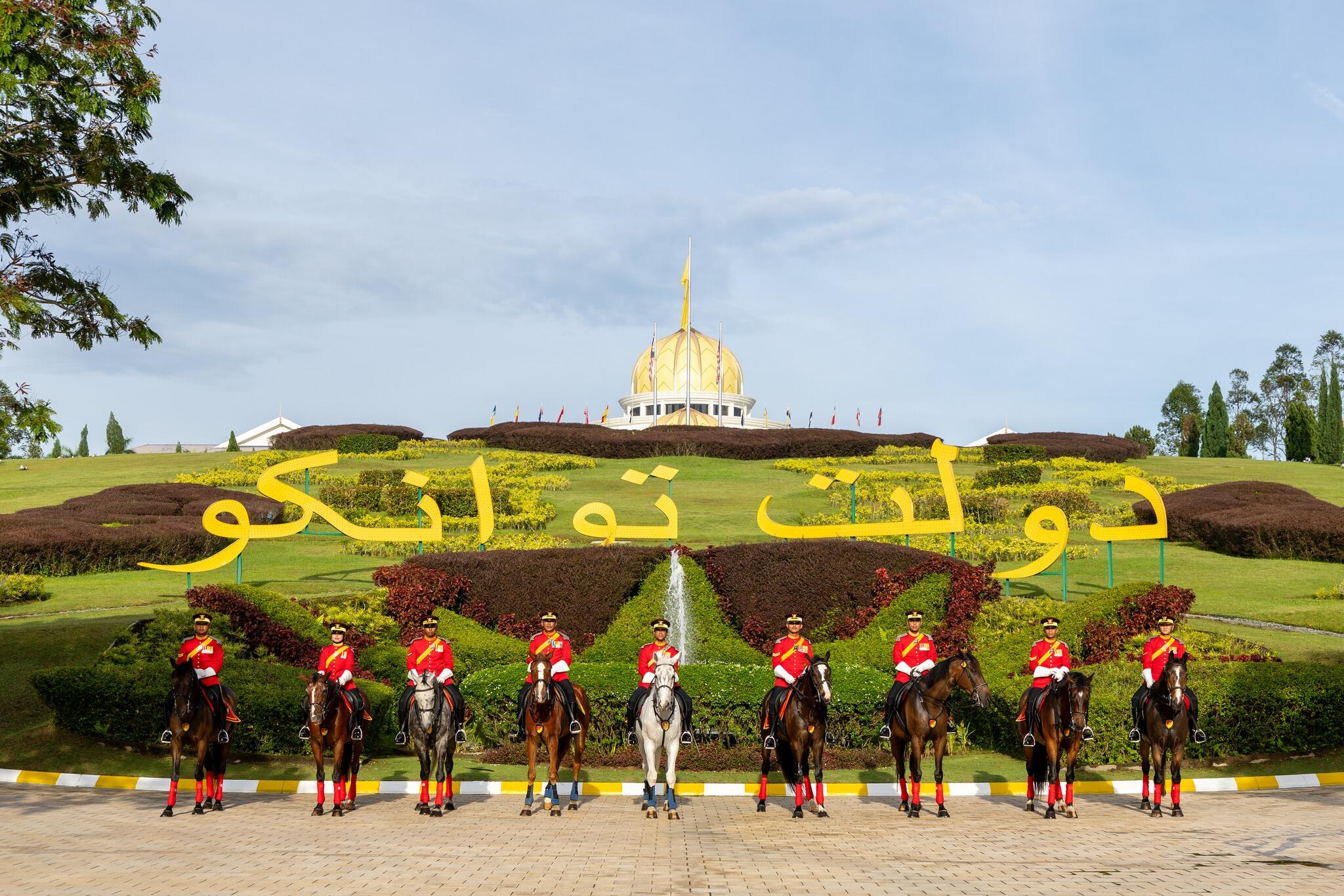 A group of mounted guards in red uniforms stand on horseback in front of Istana Negara and large text written in Arabic.