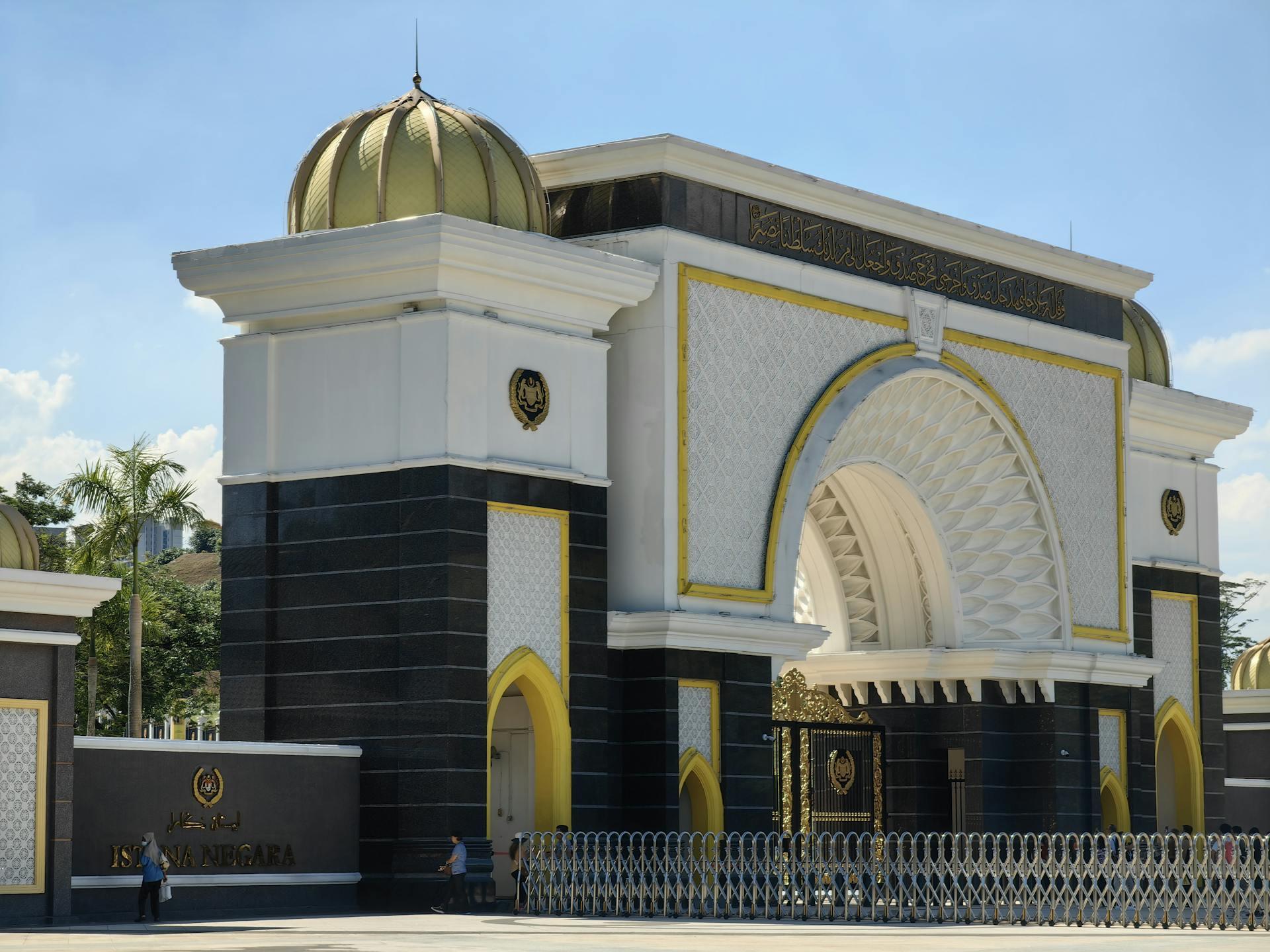 Grand entrance gate of Istana Negara in Malaysia featuring a white and black facade with gold accents, a large central Islamic arch, ornate geometric patterns, Arabic calligraphy along the top, and a prominent gold dome under a clear blue sky, with security gates and pedestrians in the foreground.