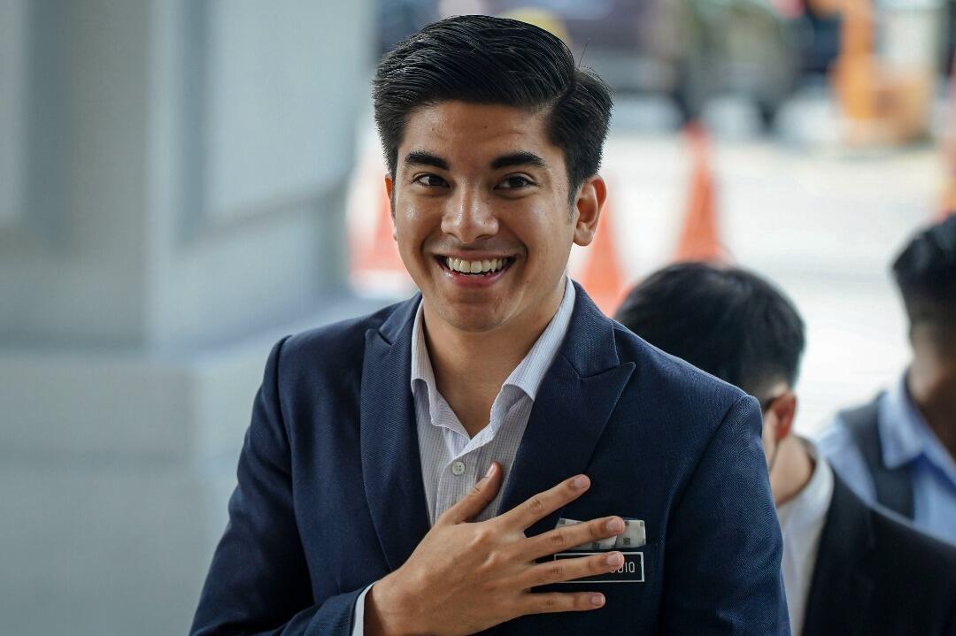 Syed Saddiq in a blue suit, walking and smiling at the camera.