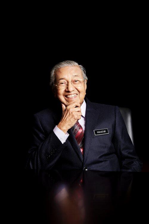 Tun Dr Mahathir Mohamad resting his chin in one hand, seated on a chair and smiling at the camera.