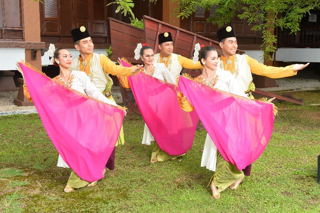 Joget Melayu - A group of dancers in traditional attire performs with bright pink scarves, exuding joy and harmony. The background features a rustic wooden building.