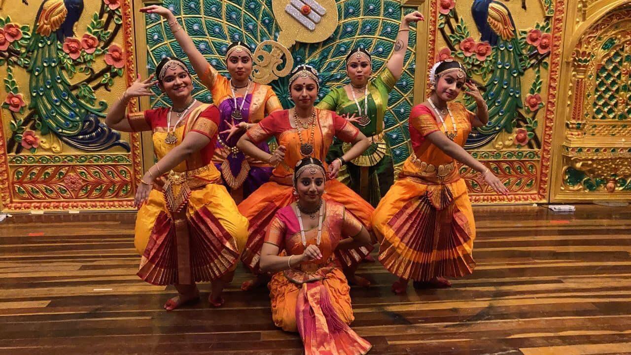 Six dancers in traditional Indian attire pose gracefully on a wooden stage against a vibrant backdrop featuring intricate peacock designs.