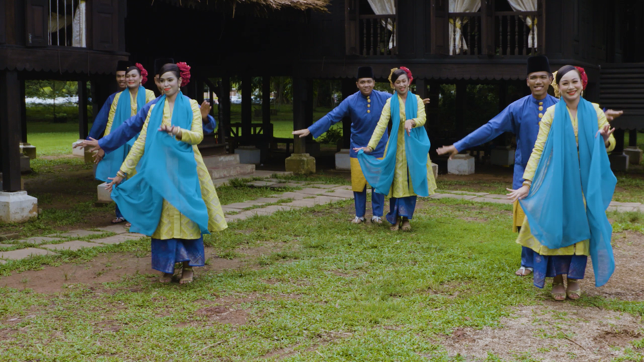 Mak Inang Dance- A group of people in traditional blue and yellow attire perform a cultural dance on a grassy area, exuding joy and harmony in front of a wooden building.