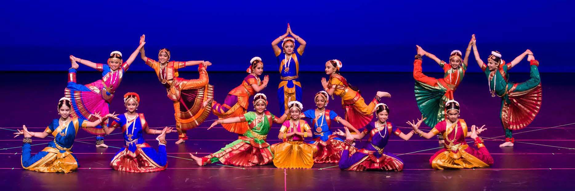 A group of young dancers in traditional Indian attire performs Bharatanatyam poses on stage, showcasing vibrant costumes and intricate movements.