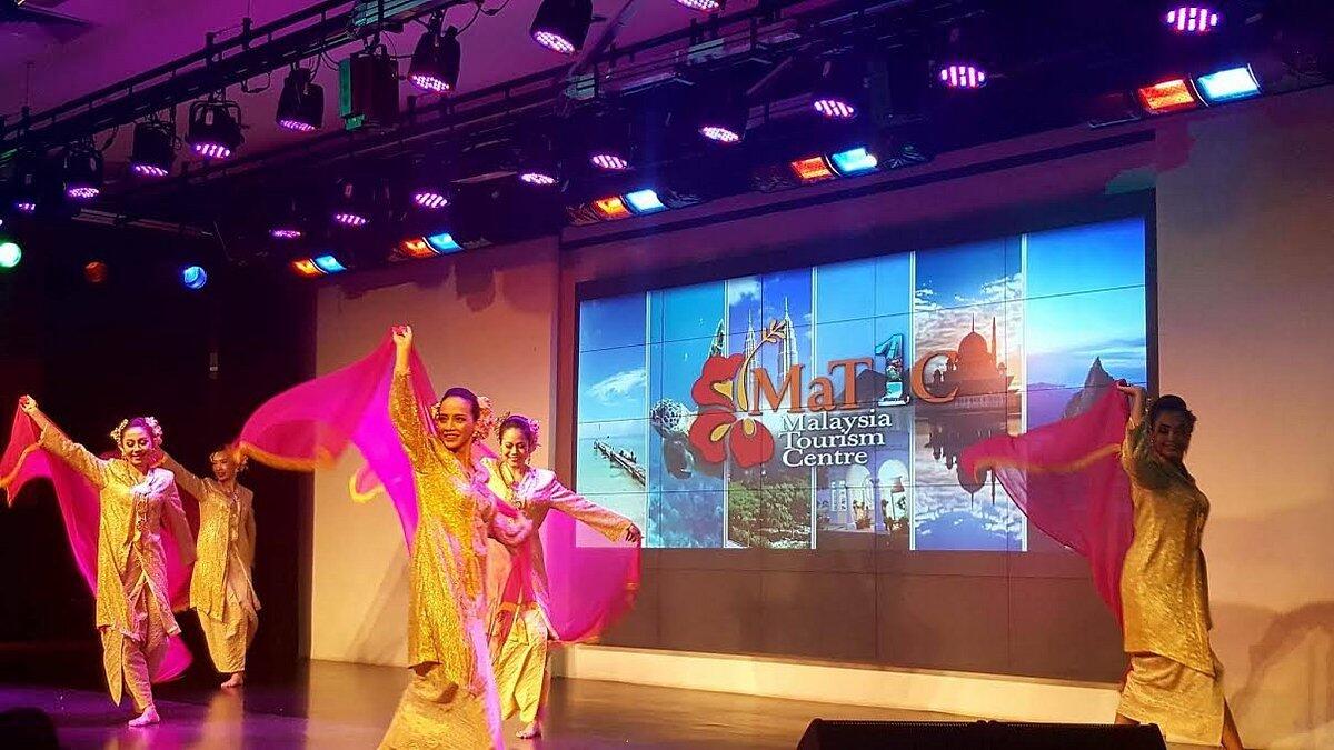 Five dancers in gold costumes with pink veils perform energetically on stage. A screen displays "Malaysia Tourism Centre" with scenic images.