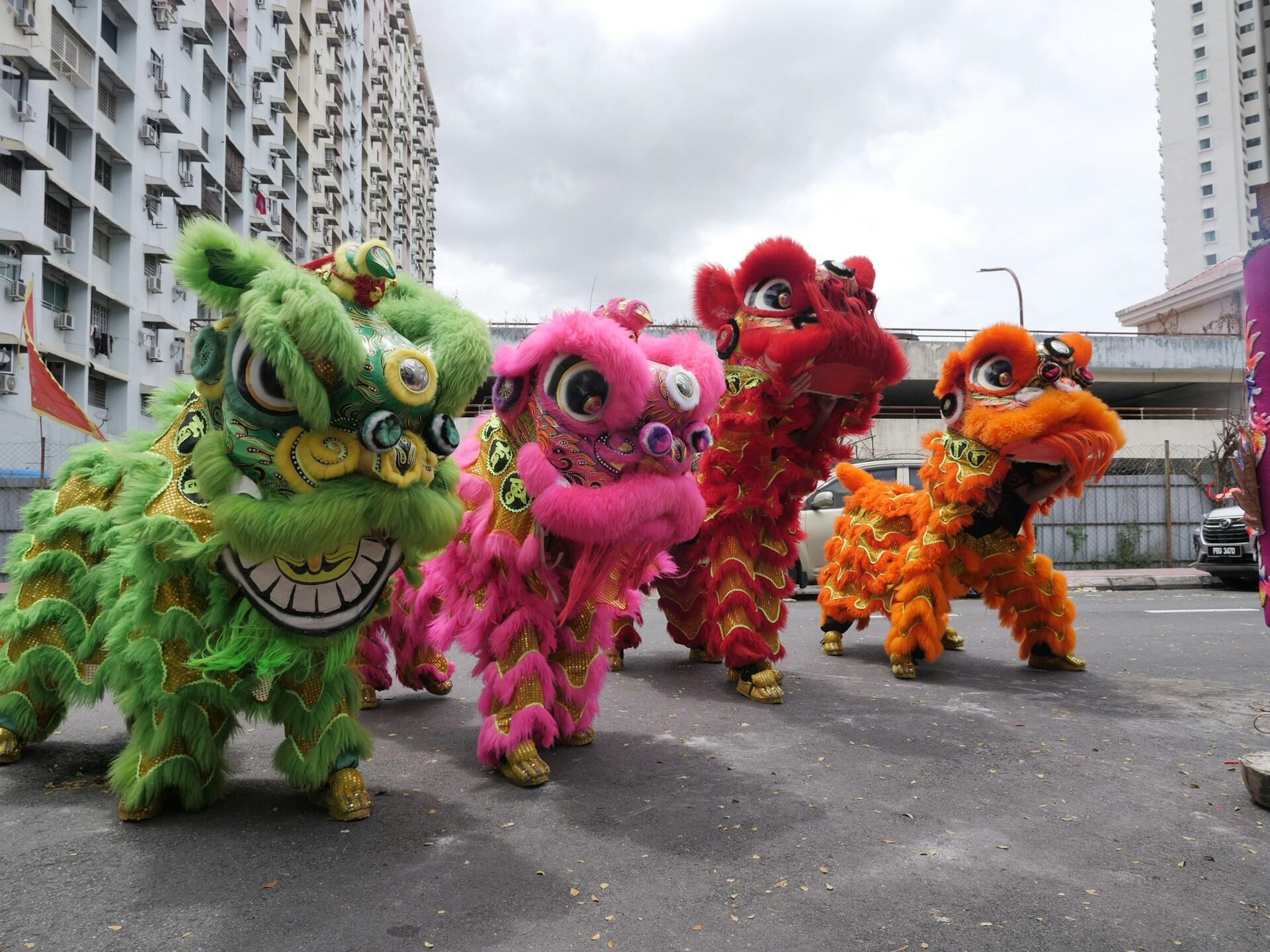 Four vibrant lion dance costumes—green, pink, red, and orange—animate an outdoor urban setting, evoking festivity and tradition, with cloudy skies above.