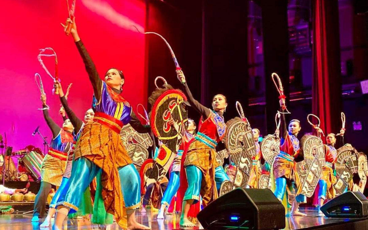 Kuda Kepang dancers in colorful traditional attire perform energetically on stage, wielding ornamental shields and whips against a vibrant red backdrop.