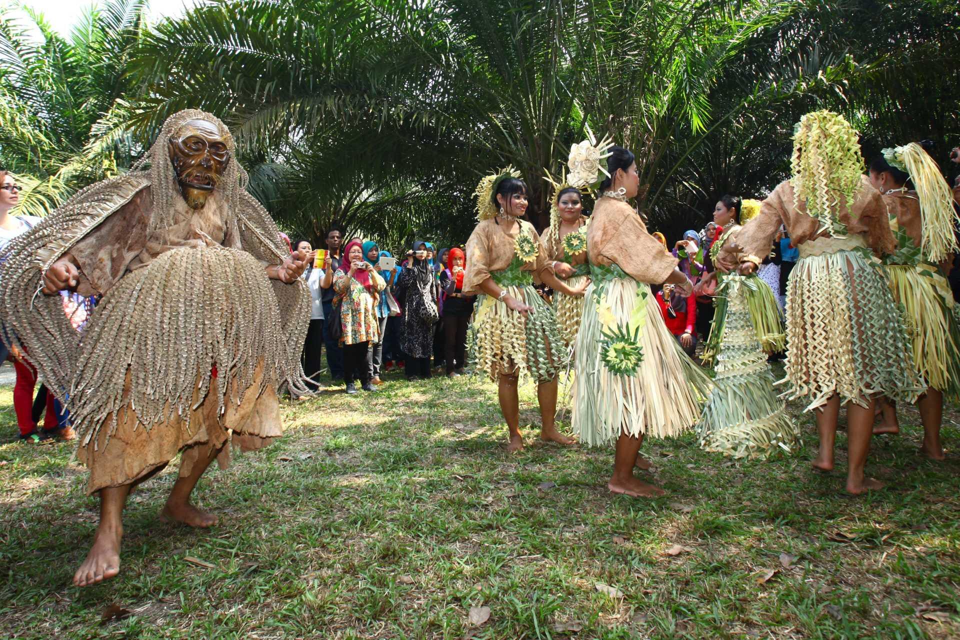 A traditional dance performance in the Mah Meri Cultural Village with dancers in ornate grass costumes. A person in an intricate mask engages in the foreground, watched by onlookers.