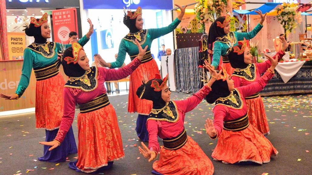 Seven women in traditional colorful attire and headpieces perform a synchronized dance at an indoor event. Their movements are expressive and graceful.