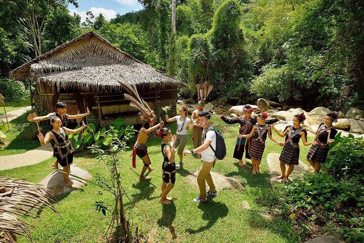 A group of people in traditional attire dance joyfully in a circle on lush greenery, near a thatched-roof hut surrounded by dense tropical foliage. Mari Mari Cultural Village.