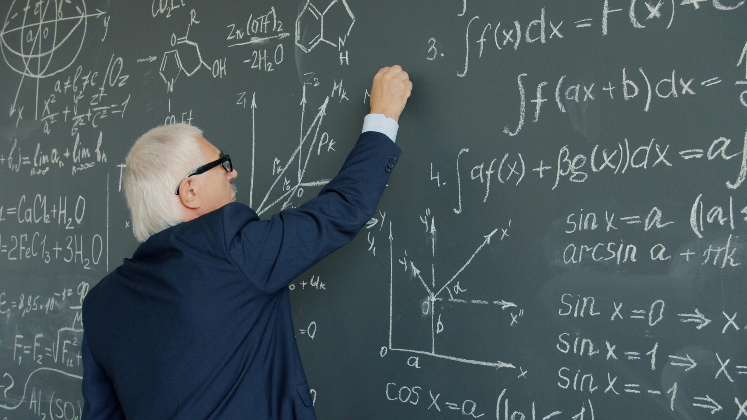 An elderly man in formal attire writes on a blackboard, engaged in a teaching or instructional activity.