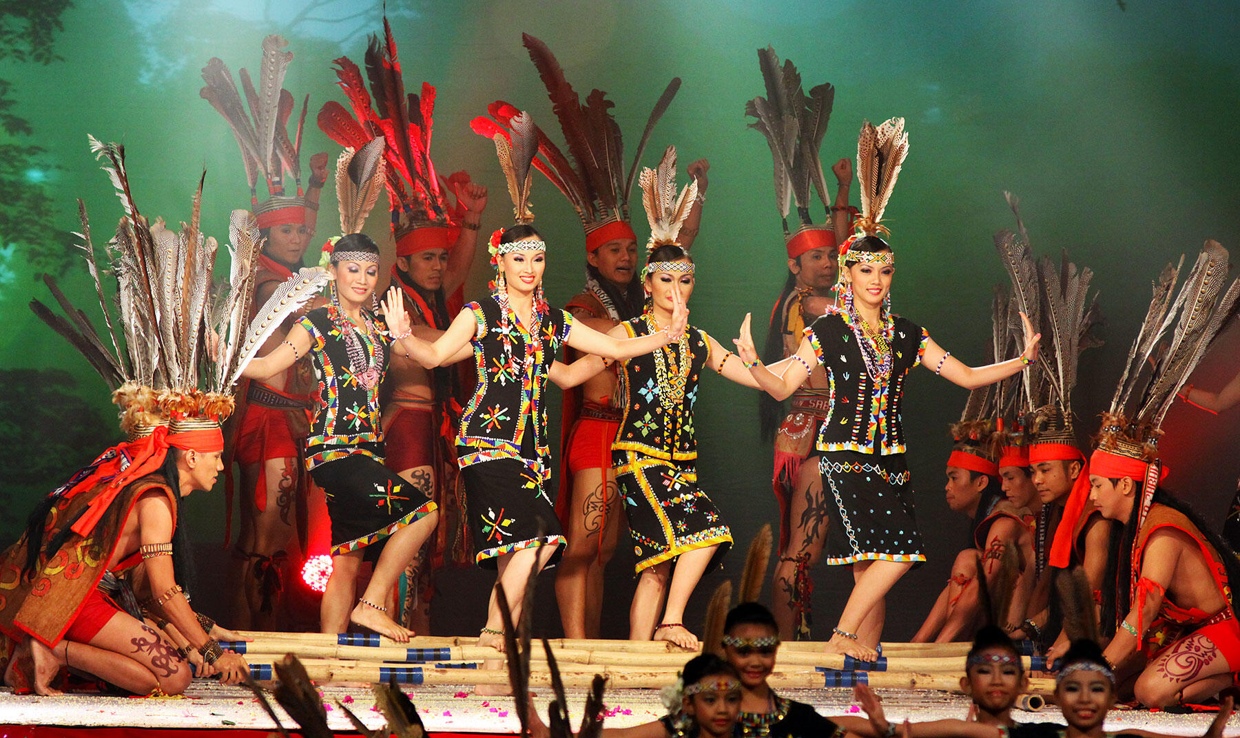 Traditional dancers perform the Sumazau dance in colourful attire and feathered headdresses on stage against a vibrant backdrop, expressing joy and cultural pride.