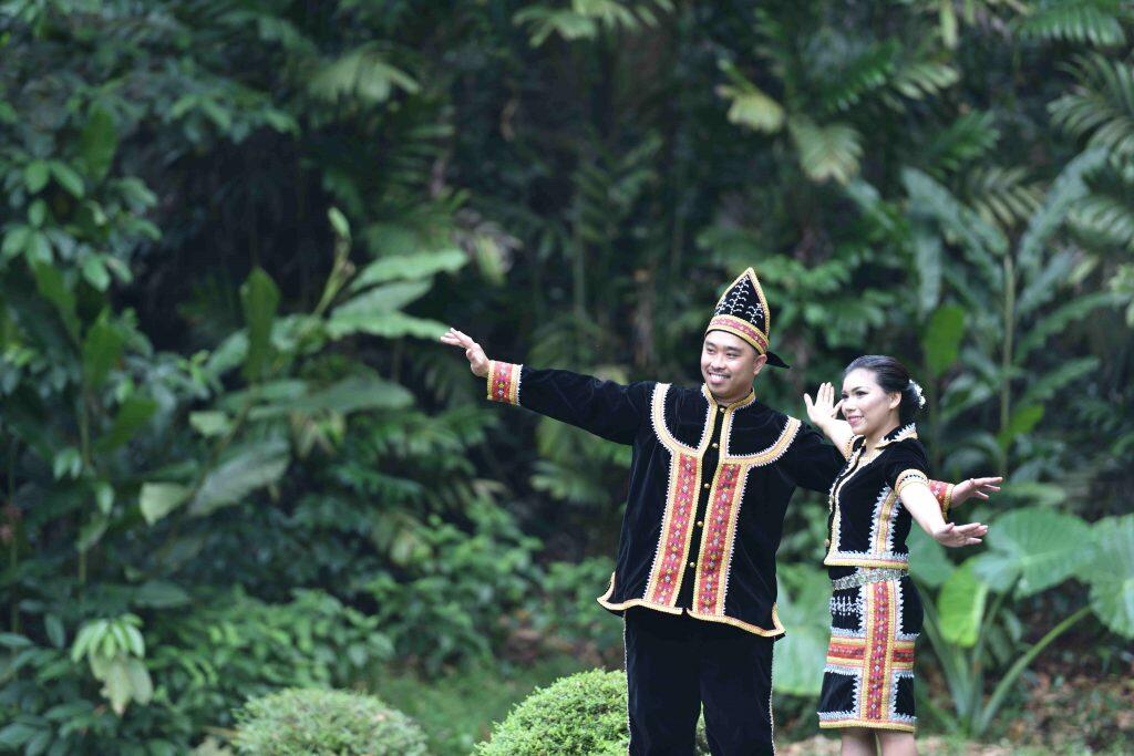 A man and woman in sabah Kadasandusun traditional attire. They are surrounded by lush green foliage, creating a vibrant, joyful atmosphere.
