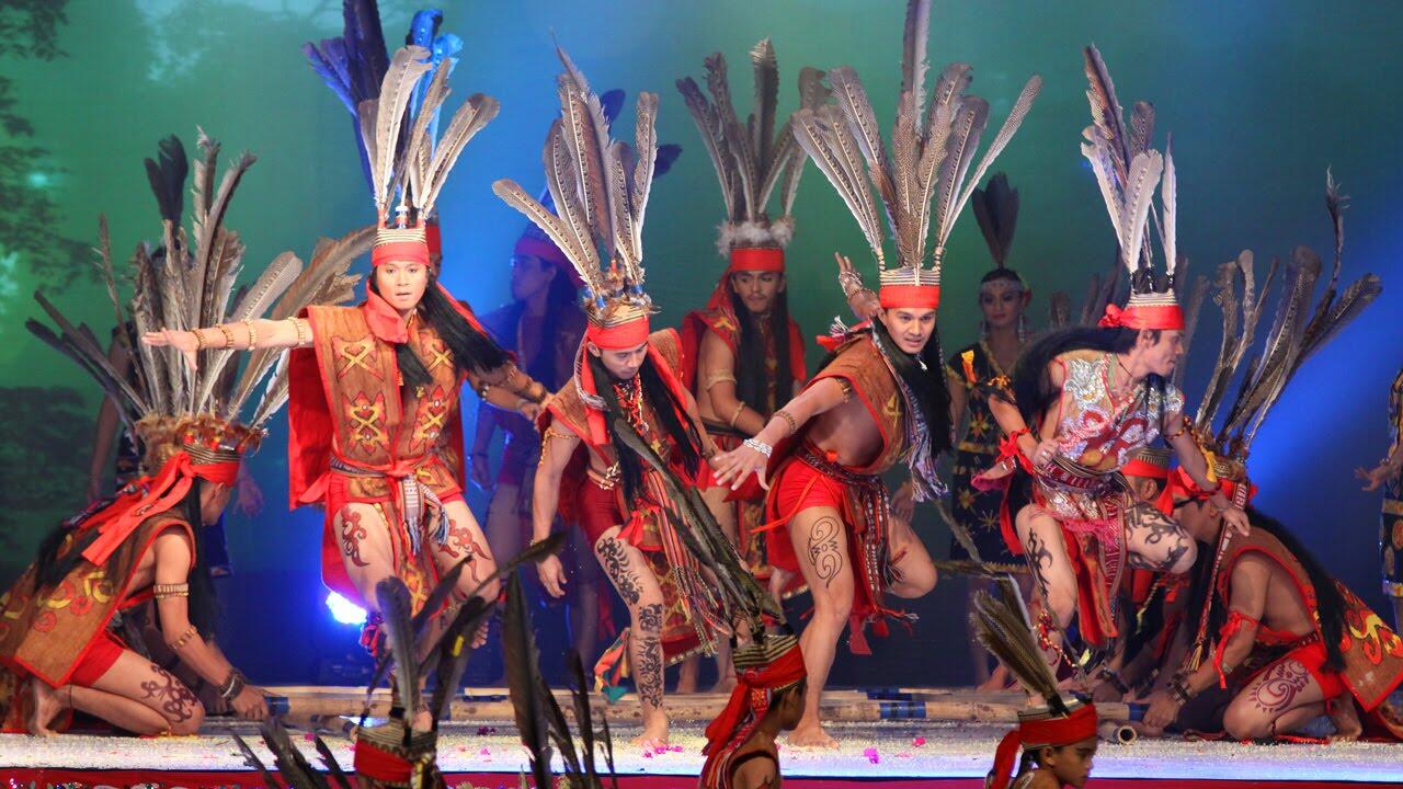 Magunatip Traditional dancers in vibrant red outfits and feathered headdresses perform energetically on stage. The scene is lively and culturally rich.ce 