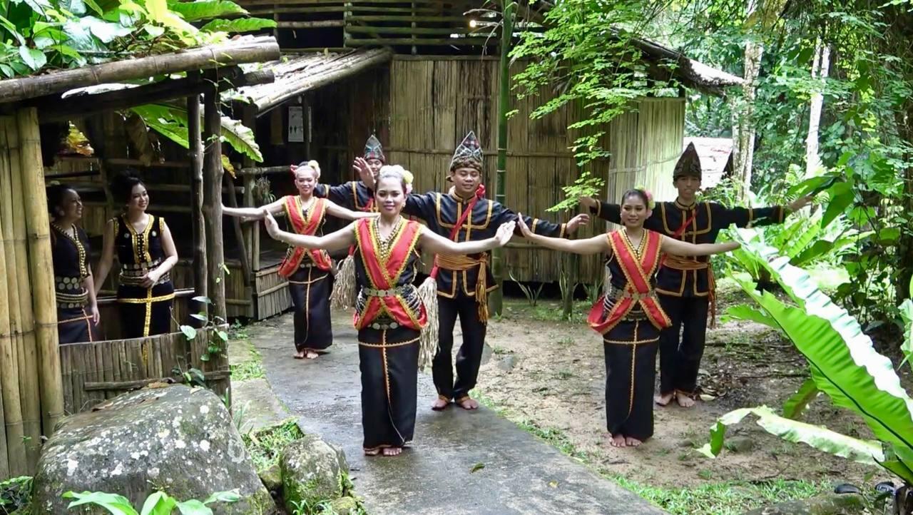 Sumazau dancers in traditional attire perform a cultural dance in a lush, tropical setting. Their expressions are focused and joyful, conveying unity.