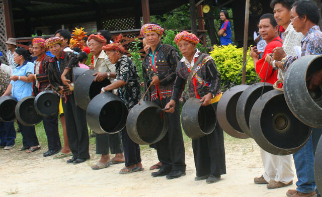 A row of traditionally dressed individuals plays large gongs in an outdoor setting, conveying a sense of cultural celebration and community.