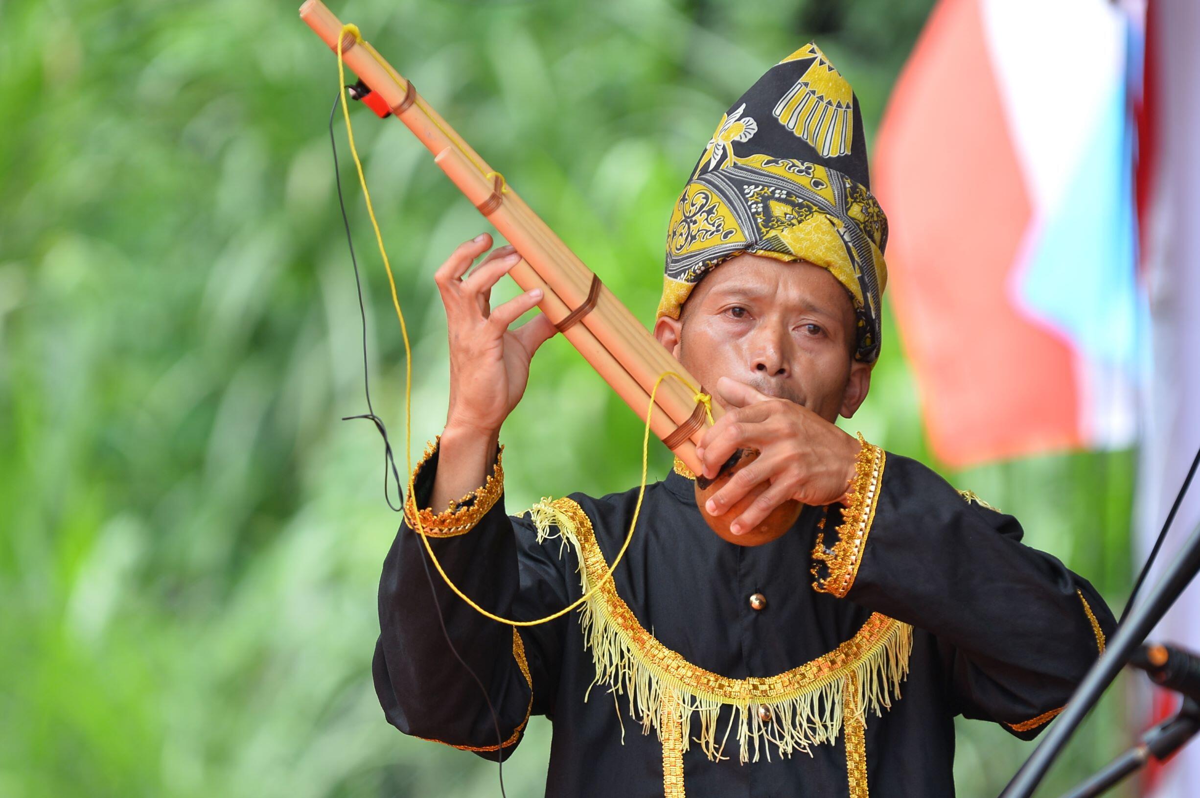 A man in traditional attire plays a bamboo flute (Sompoton), wearing an ornate black and yellow headdress. He stands against a backdrop of greenery and blurred flags.