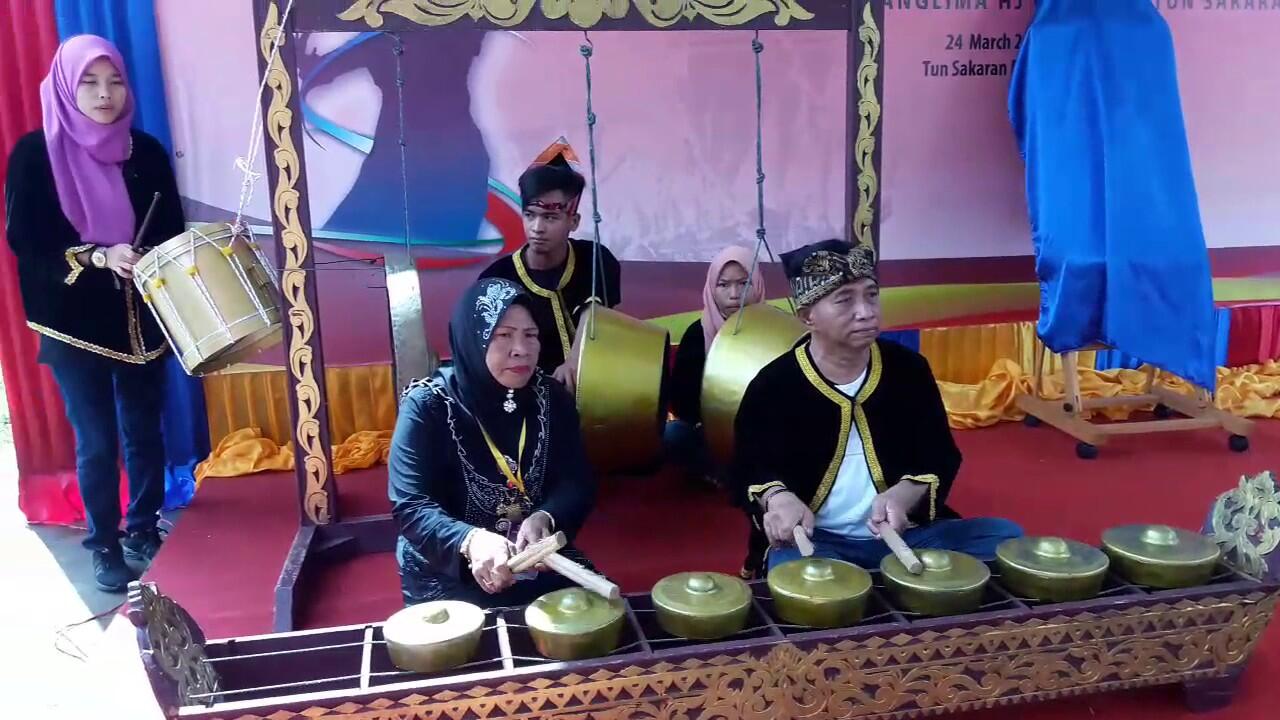 A group of people in traditional attire perform with a gong ensemble on a stage. The mood is focused and cultural, featuring golden instruments.