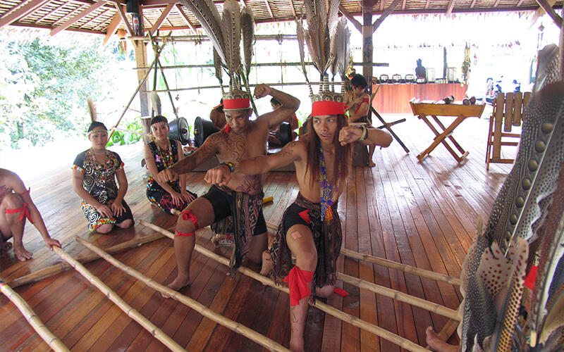 Magunatip dance group in traditional attire perform a lively cultural dance inside a wooden pavilion. They are energetically posing with expressive gestures.