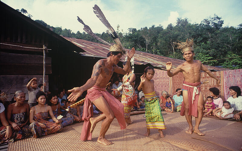 Two men and a child in traditional attire perform the Ngajat dance, surrounded by a seated audience. The scene is vibrant, lively, and cultural.