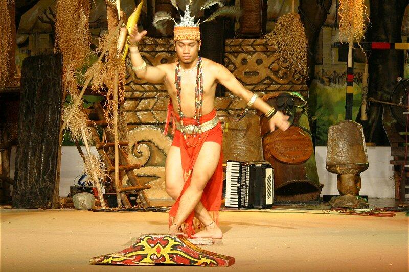 A traditional Ngajat dancer in vibrant attire performs energetically on stage, surrounded by tribal decorations and musical instruments, conveying cultural richness.