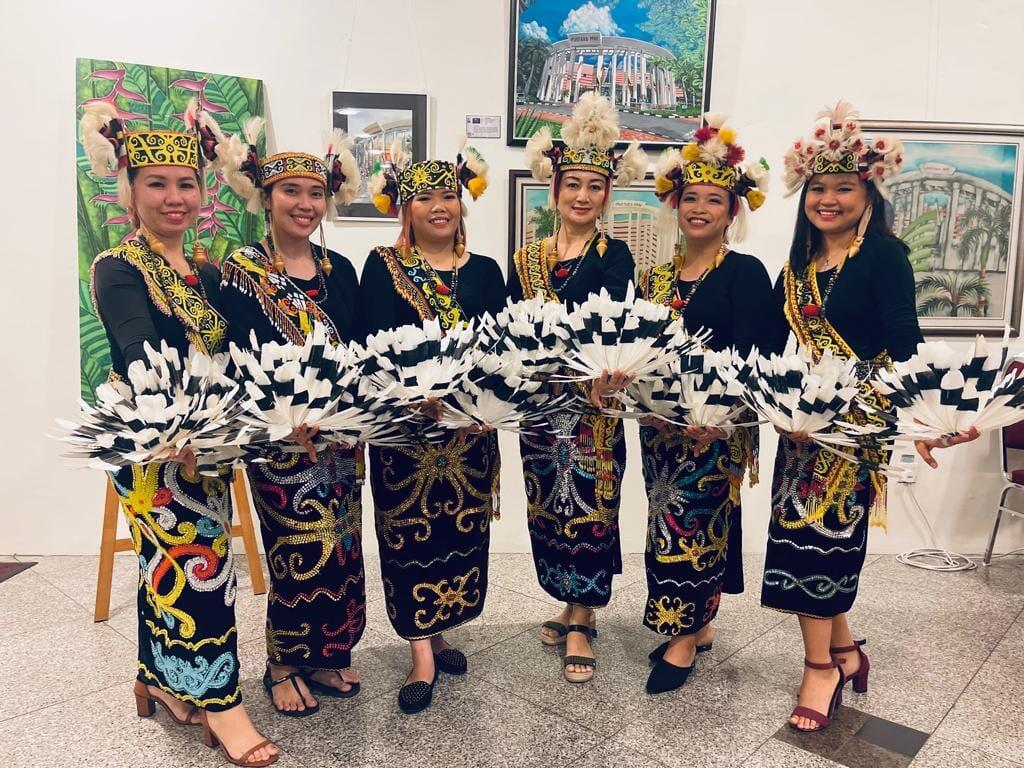Datun Julud dancers - Six women in vibrant traditional attire stand smiling, holding decorative fans. They're against an art-filled backdrop, conveying cultural pride and joy.