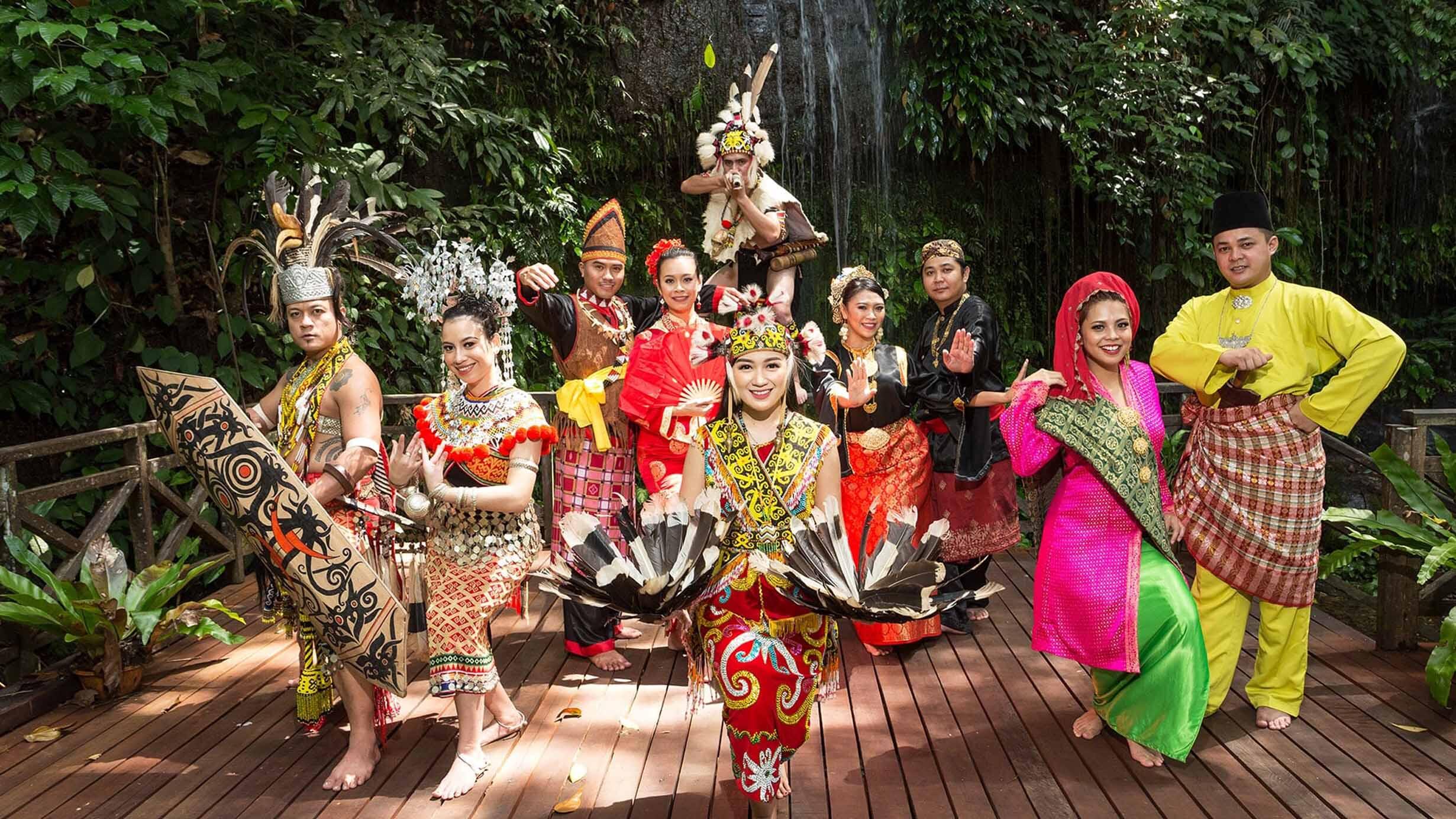 A group of people in vibrant Sarawak traditional dance attires pose joyfully on a wooden deck in a lush forest. They showcase cultural diversity and celebration.
