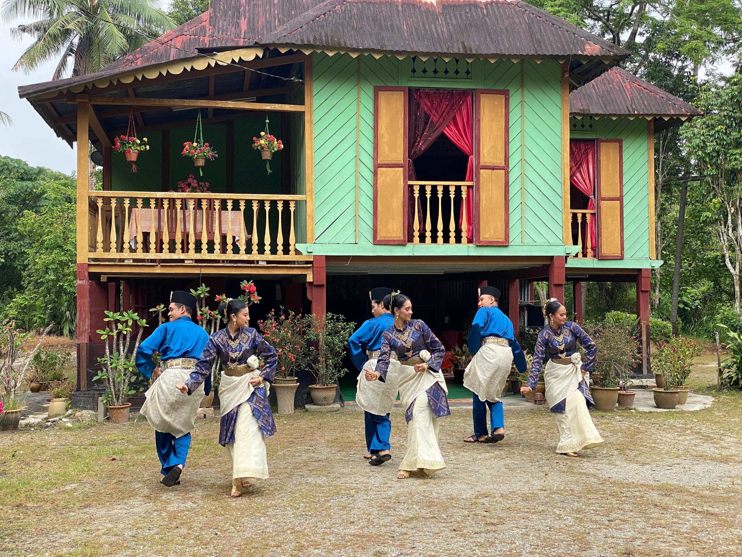 A group of Zapin dancers in traditional Malaysian attire perform a cultural dance in front of a vibrant, green wooden house with red curtains and potted plants.
