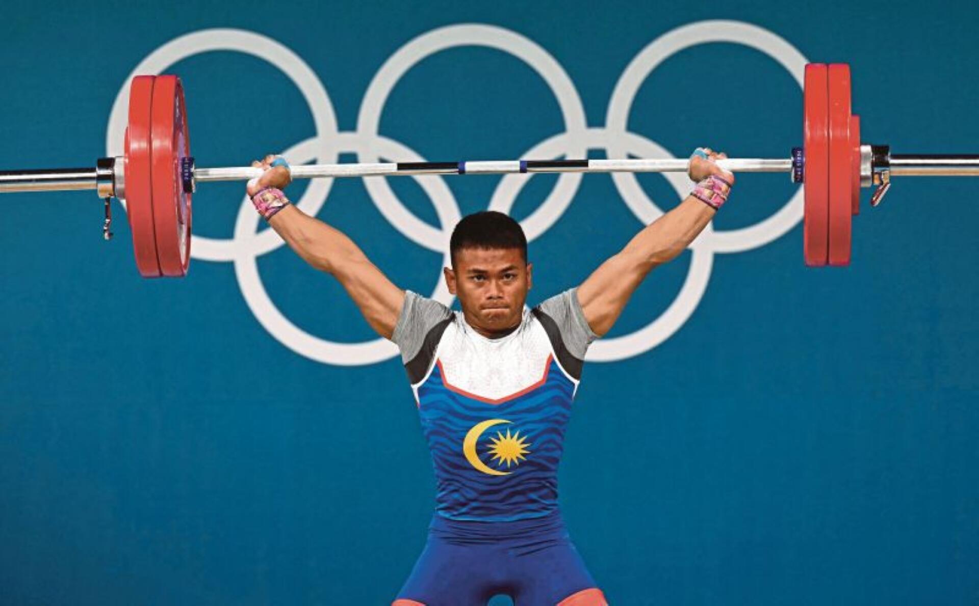 Weightlifter in blue attire with a crescent and star lifts heavy barbell at the Olympics, with Olympic rings in the background.
