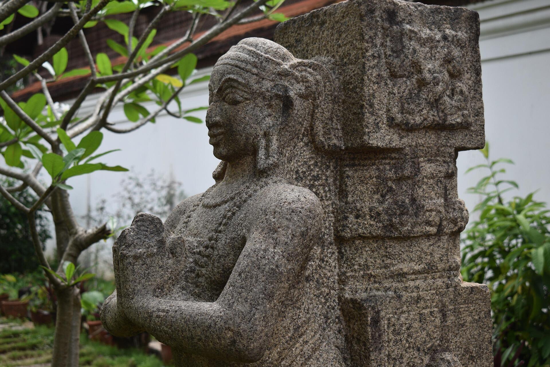 Ancient stone sculpture of a Tamil figure standing beside a carved pillar in a lush garden setting.
