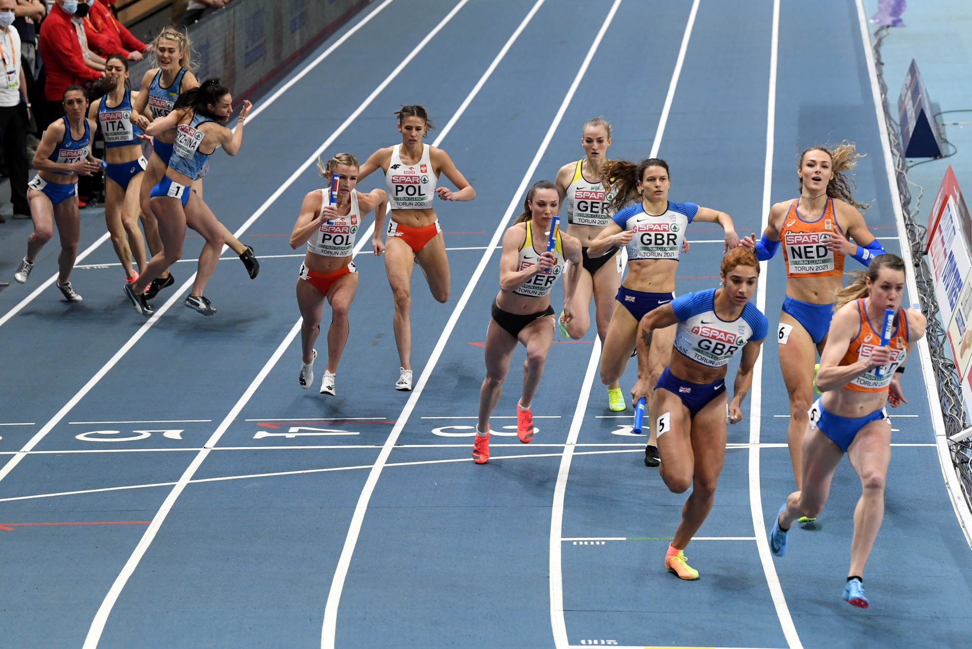Female athletes from multiple countries passing the baton during a relay race on an indoor track.