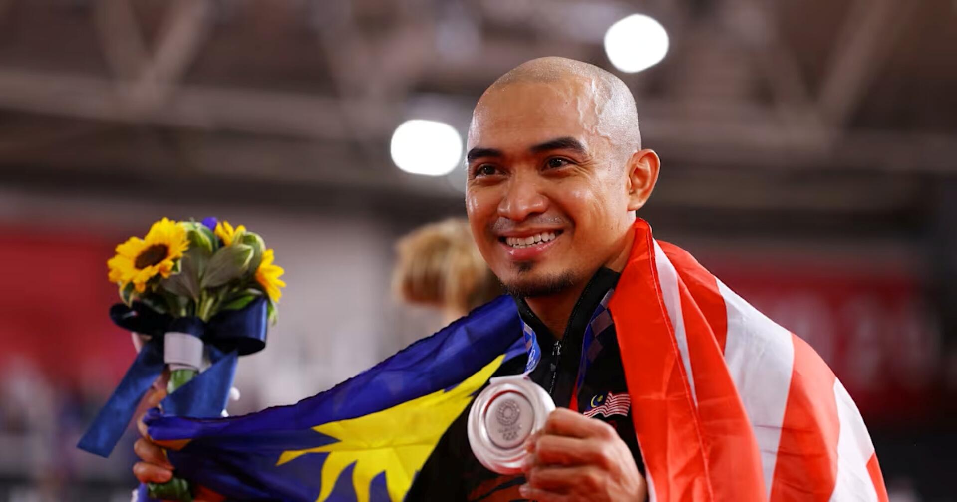 A smiling athlete draped in a Malaysian flag holds a silver medal and a bouquet of sunflowers, conveying pride and achievement in a brightly lit venue.