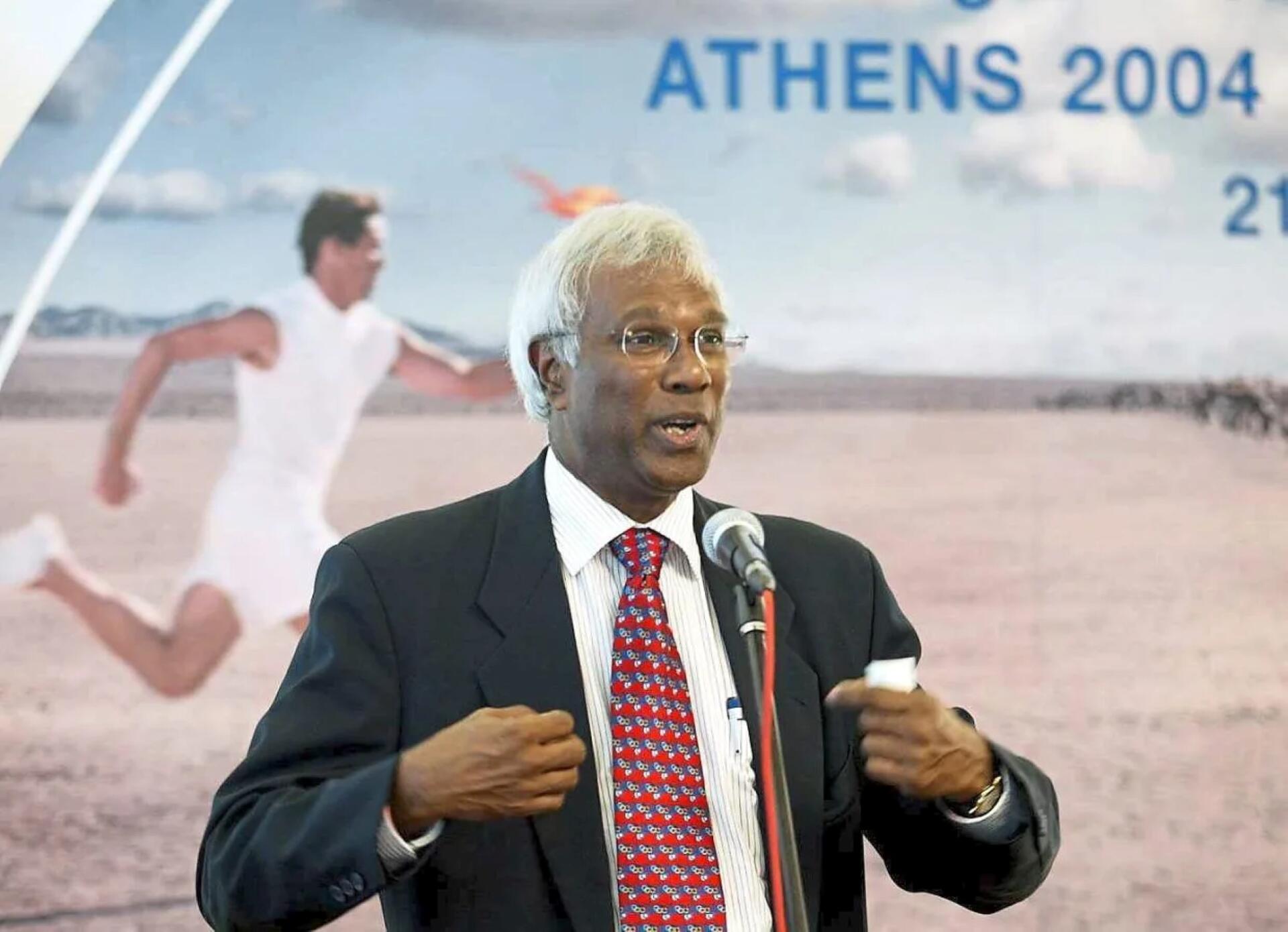 Elderly man with white hair and glasses speaks at an event, wearing a suit and red tie. Behind him, a poster shows an Olympic runner and "Athens 2004."