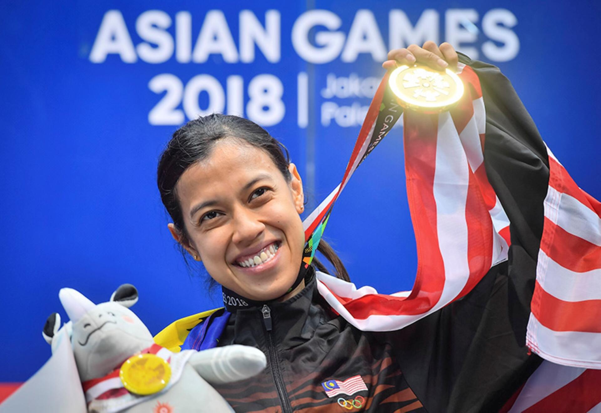 Female athlete smiling, holding a gold medal and wrapped in a flag at the Asian Games 2018. A plush toy is in the foreground. Jubilant mood.