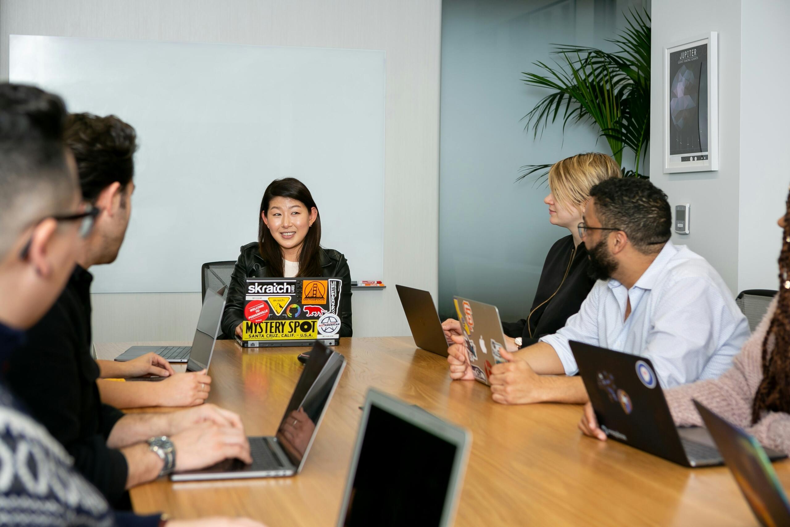 A group of five individuals seated at a table, each using laptops, engaged in a collaborative discussion.