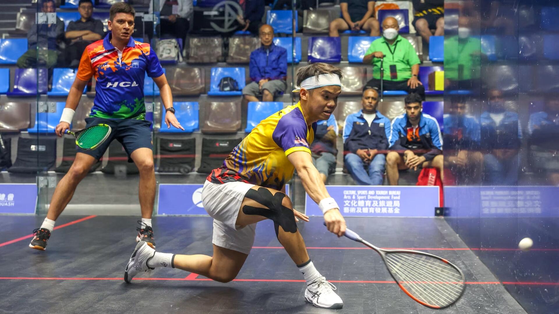 Two male squash players in action on a squash court with spectators seated in the background.