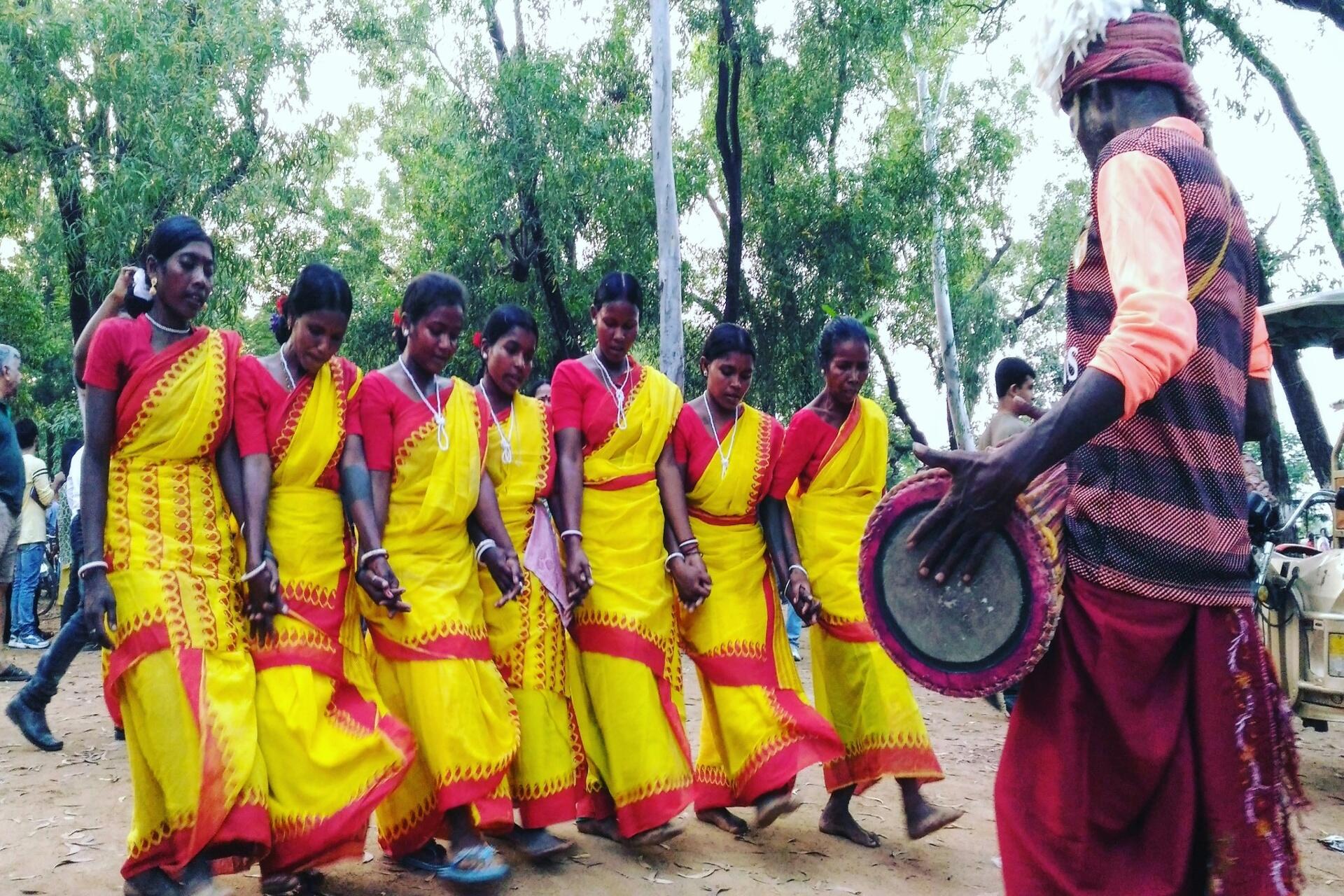 Women performing a traditional folk dance in colourful attire while a drummer plays music during a cultural celebration.