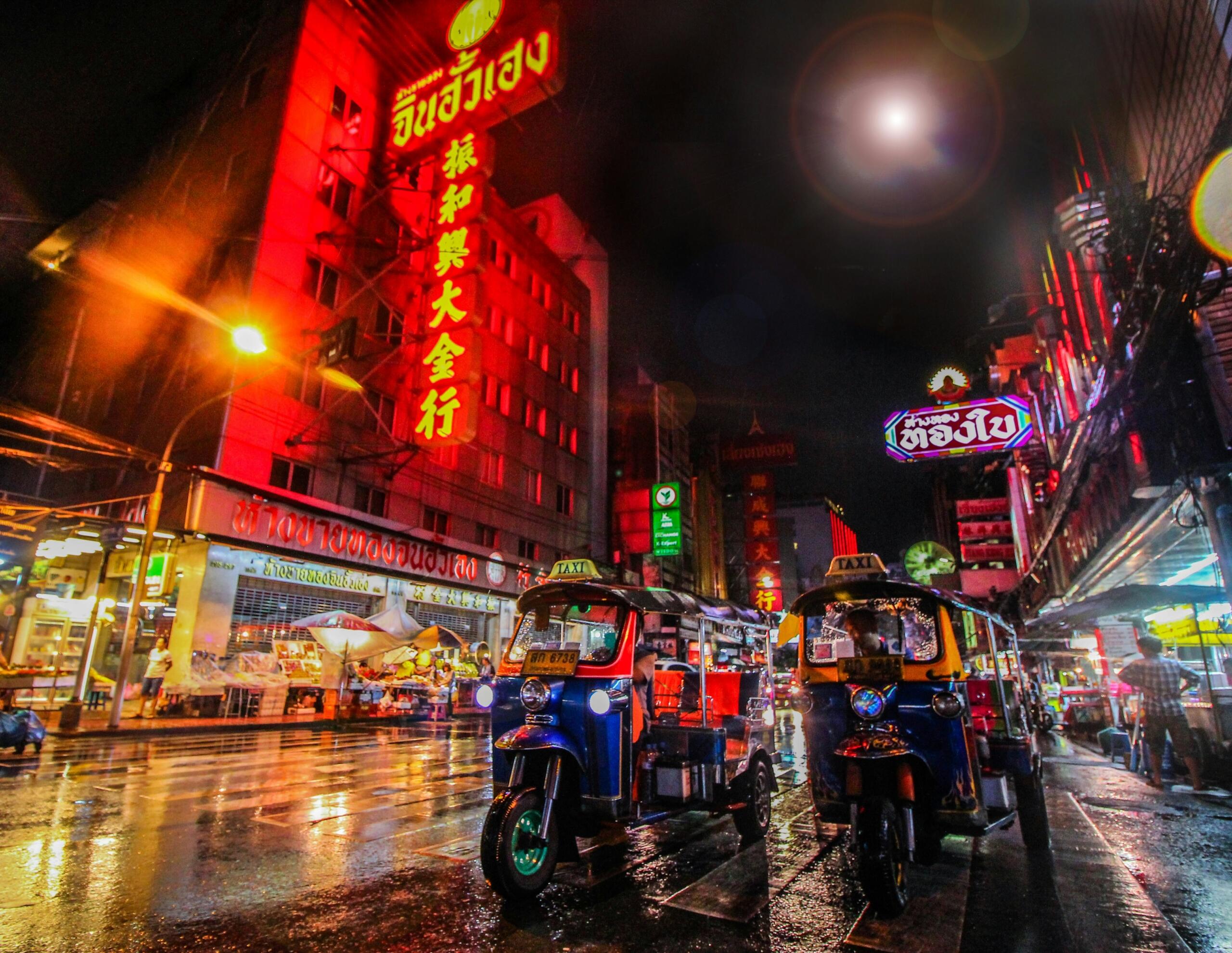 A street scene featuring two parked auto rickshaws beside a vibrant neon sign illuminating the surroundings.